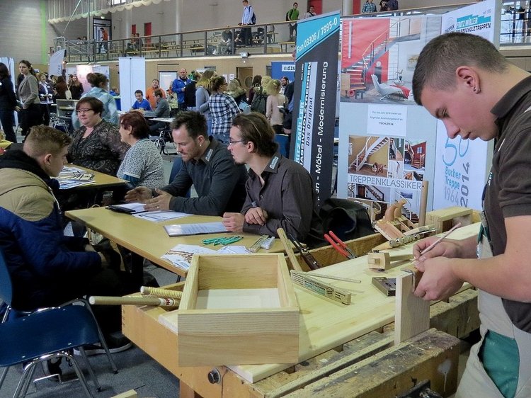 Das Foto zeigt den Messestand der Firma Fritz Müller, drei Personen sind im Gespräch, ein junger Mann bearbeitet Holzteile