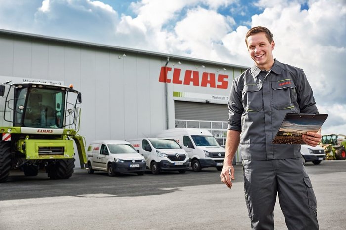Das Foto zeigt einen jungen Mann in Atbeitsbekleidung vor einer CLAAS-Maschinenhalle mit davor parkenden Kraftfahrzeugen