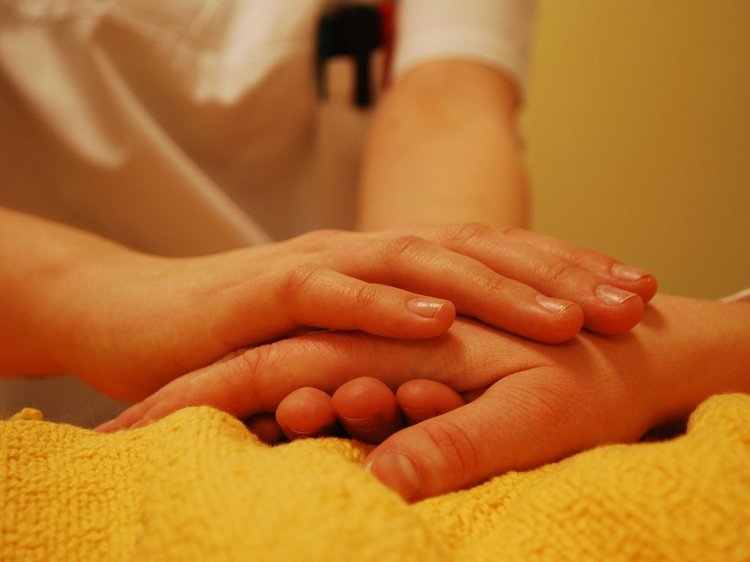 Das Foto zeigt stellvertretend für alle pflegenden und medizinische Berufe zwei Hände zweier Personen auf dem Krankenbett