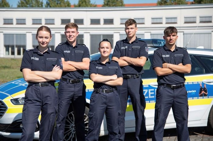 Das Foto zeigt vier Polizisiten und eine Polizisitin in Uniform vor einem Polizeiauto