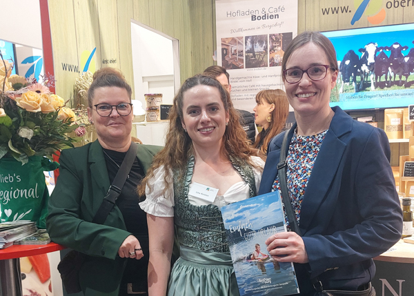 Auf dem Bild sind zu sehen stehend von links nach rechts: Kerstin Tammer, (Touristinfo Fürstenberg/Havel), Lina Kersten im Dirndel (Mitarbeiterin vom Landkreis Oberhavel) und Victoria Czinczel (Touristinfo Stechlin). Alle drei stehen vor einem Messestand in einer Messehalle. Frau Czinczel hält die aktuelle Ausgabe des Gasgeberverzeichnisses in die Kamera.