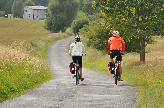 Das Foto zeigt eine Frau und einen Mann von hinten auf ihren Fahrrädern sitzend auf einem asphaltierten Radweg durch eine grüne Landschaft fahrend, links und rechts von ihnen ist Wiese, im Hintergrund ist ein graues Haus zu sehen, auf das der Weg zuführt. Der Weg ist auf beiden Seiten mit Bäumen gesäumt.